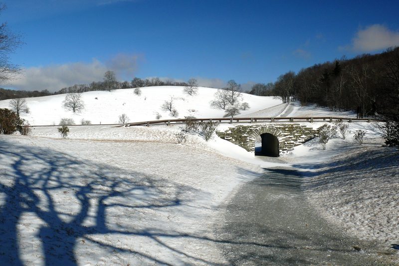 Snow-covered carriage trail in Moses H. Cone Memorial Park curving toward a stone-arched tunnel, with snowy hills and bare trees under a blue sky